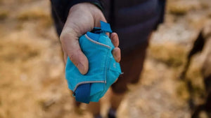 A person holding a compact, blue Ruffwear dog harness in a natural outdoor setting.